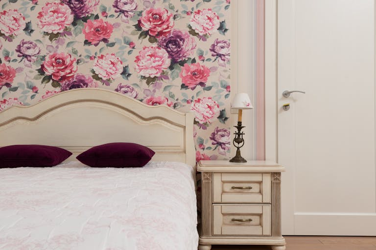 Interior of cozy classic bedroom with accent floral wall behind bed and bedside table with lamp near white doorway